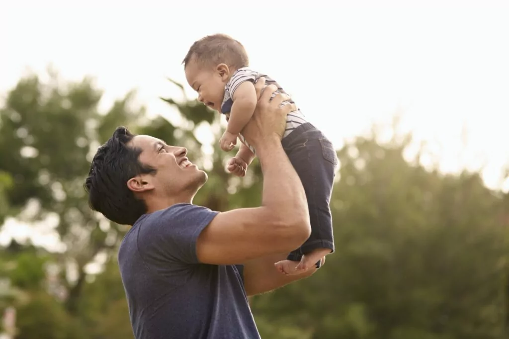 Millennial Hispanic father holding his little baby in the air in the park, close up