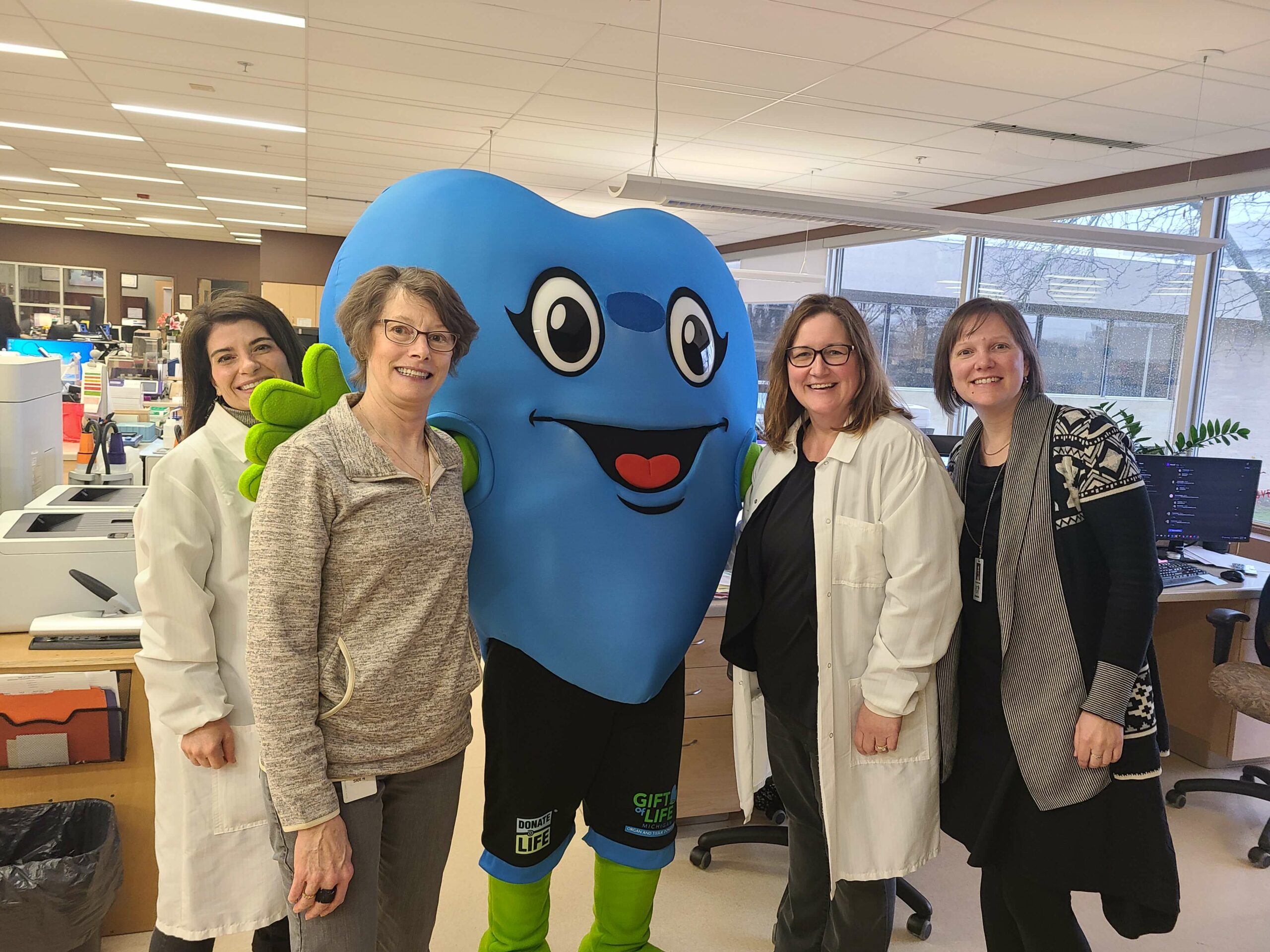 Four laboratory staff, some in white coats, stand next to Hartley, the Gift of Life Michigan mascot.