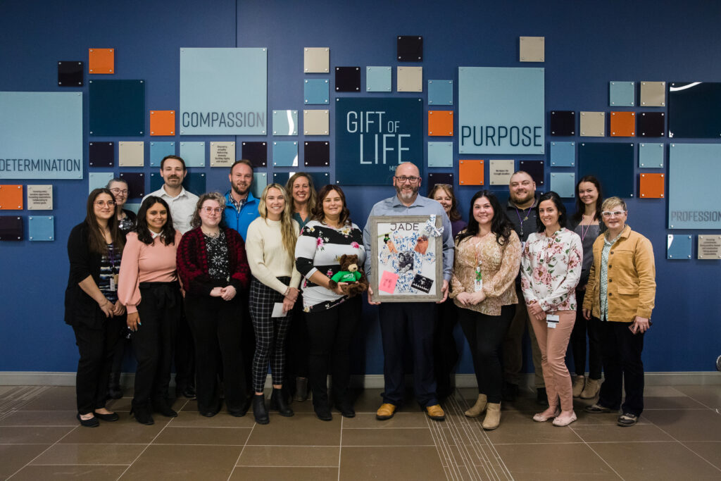 16 people standing in front of Gift of Life's core purpose display, including two parents holding photos of their infant daughter