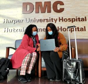 Two women wearing surgical masks, working on a laptop, sitting under a DMC Harper University Hospital sign