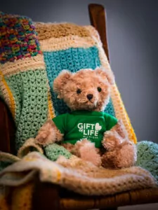 brown teddy bear wearing a green Gift of Life logo shirt sitting on an afghan blanket