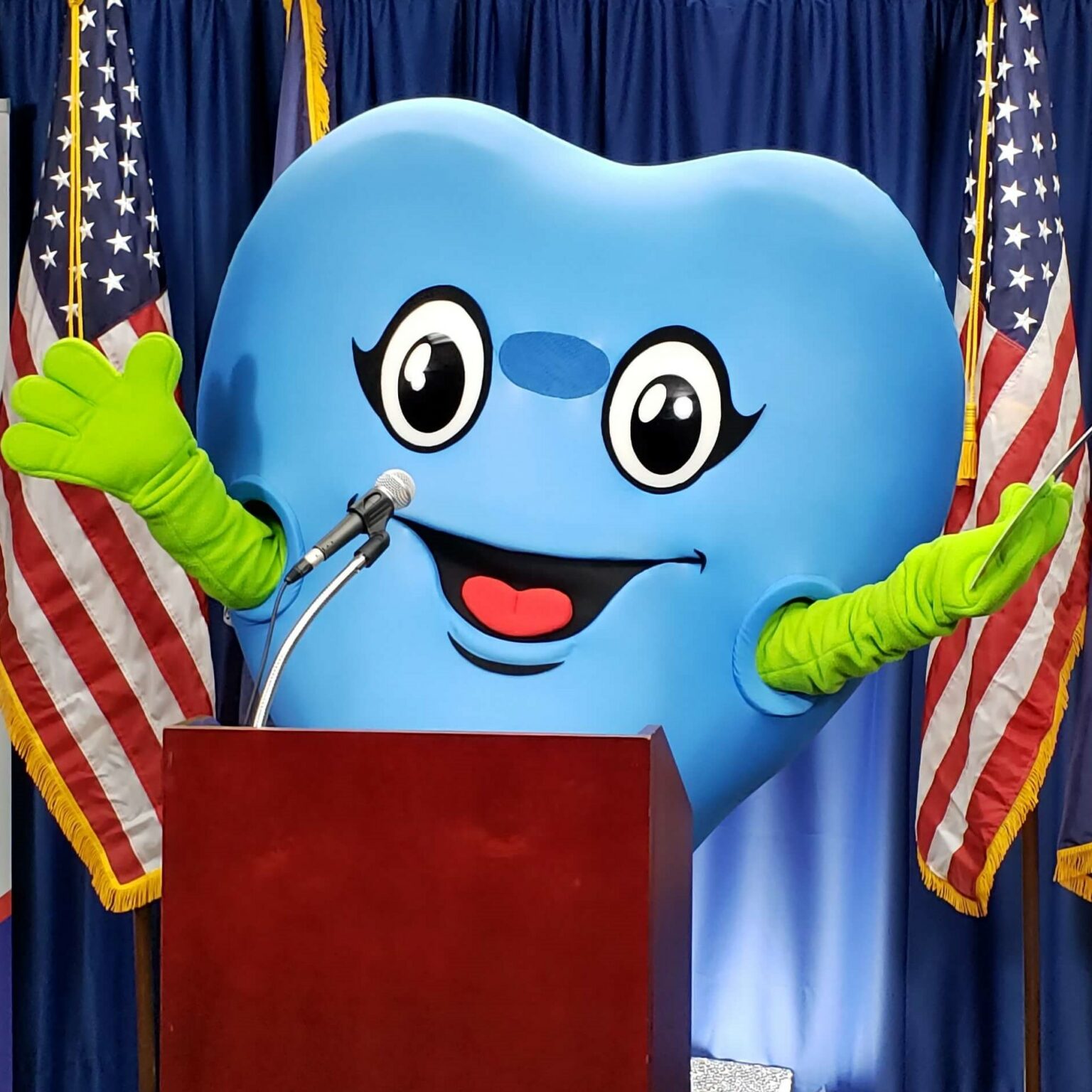 Blue heart-shaped mascot at podium with American and Michigan flags behind them