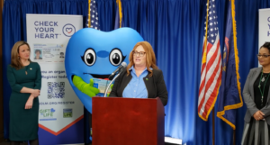 Woman at podium, blue heart shaped mascot and two other women behind her