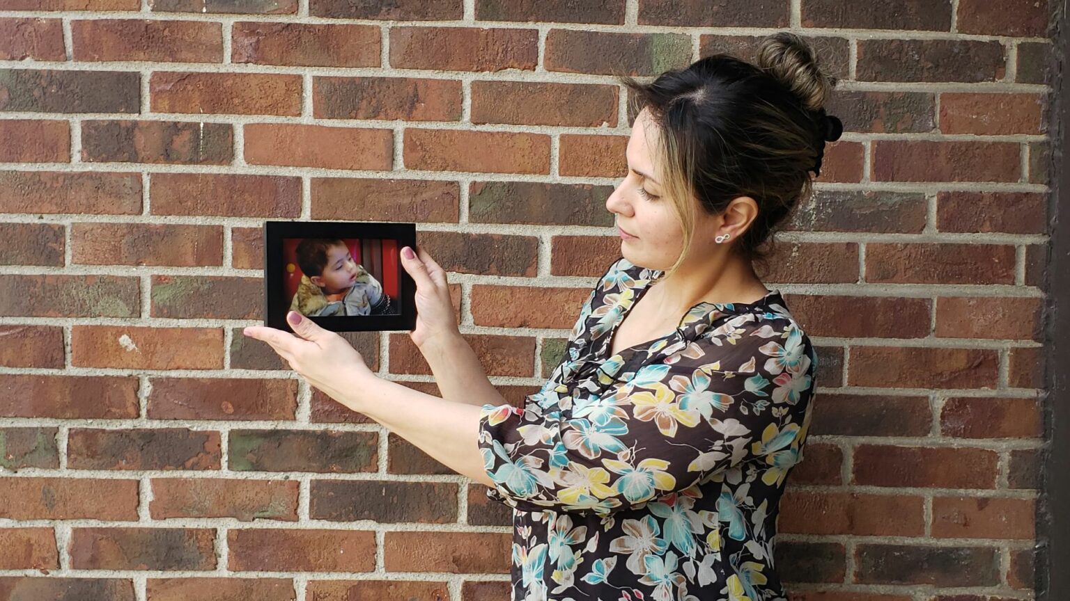 A woman standing in front of a red brick wall holding a framed photo of a young boy