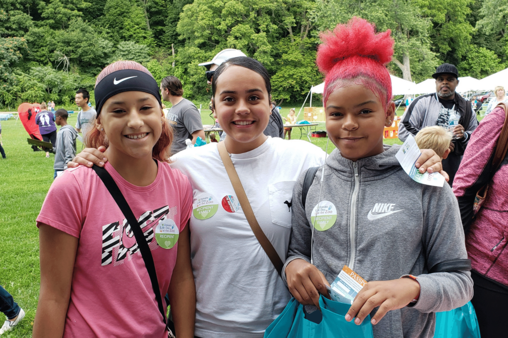 Three teen girls standing next to each other at an outdoor event with grass and trees behind them