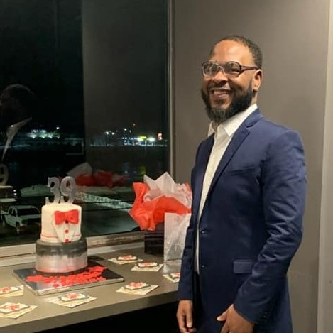 Patrick Price, a bearded Black man with glasses in a blue suit, stands in front of a birthday cake and smiles at the camera