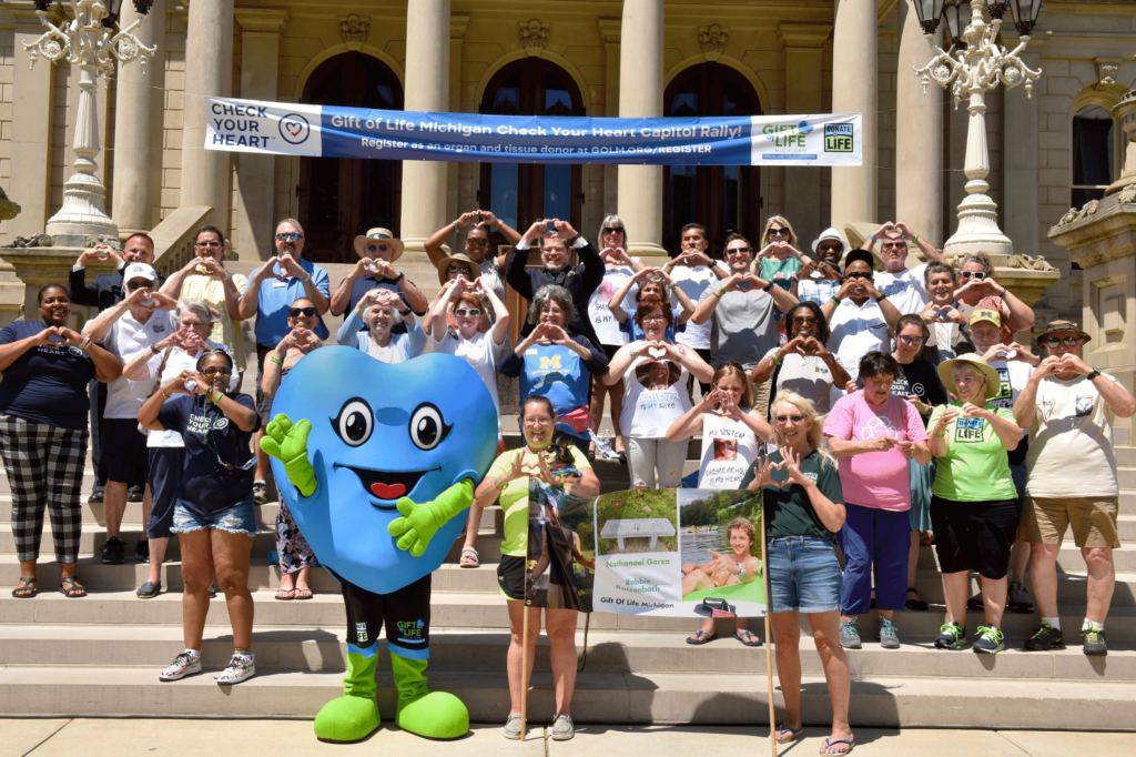 Photo of a crowd and a blue heart mascot in front of the Michigan State Capitol building, holding their hands in the shapes of hearts