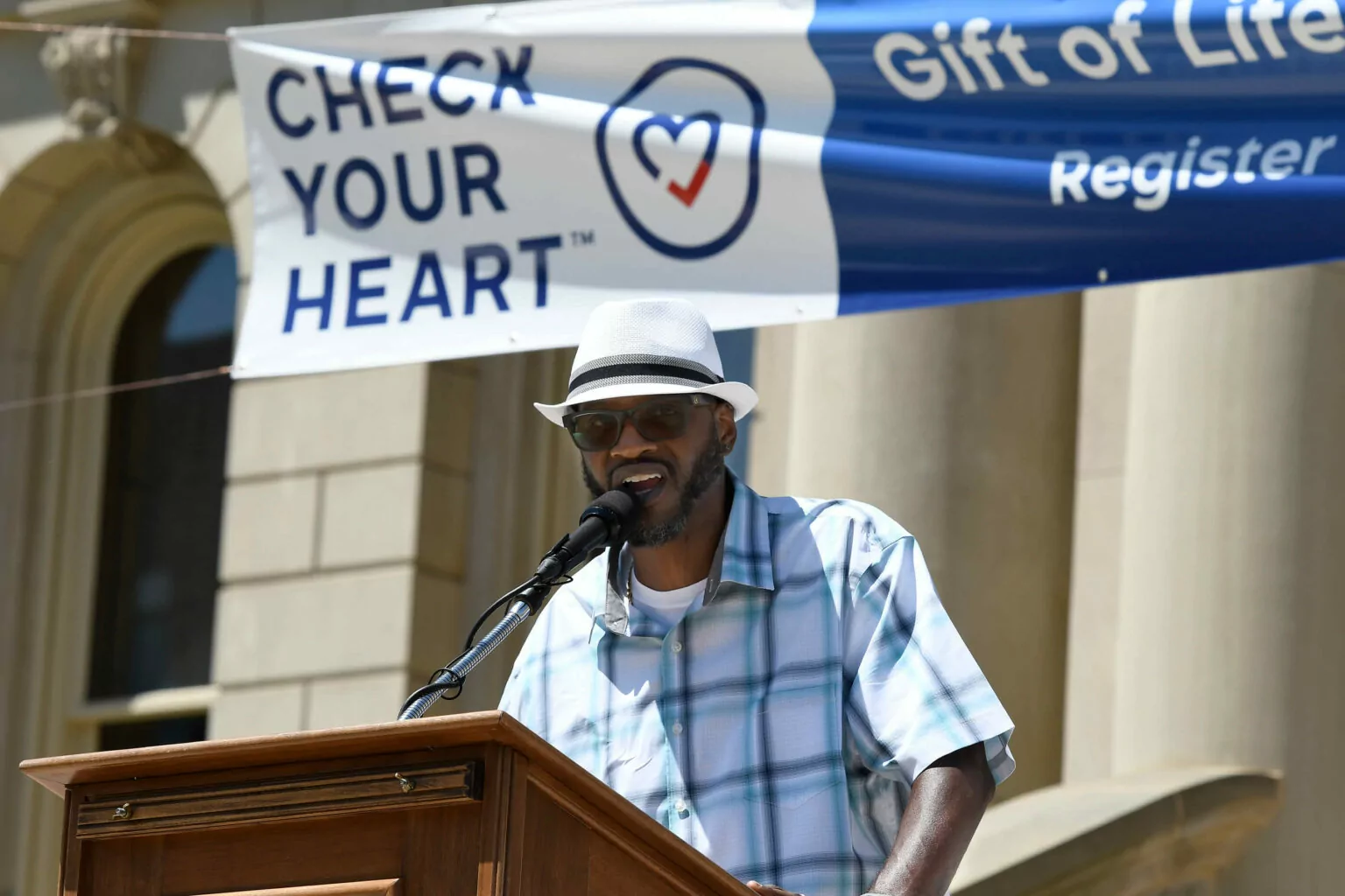 Donor father John Edmond at the podium on the Capitol Steps with a Check Your Heart banner behind him