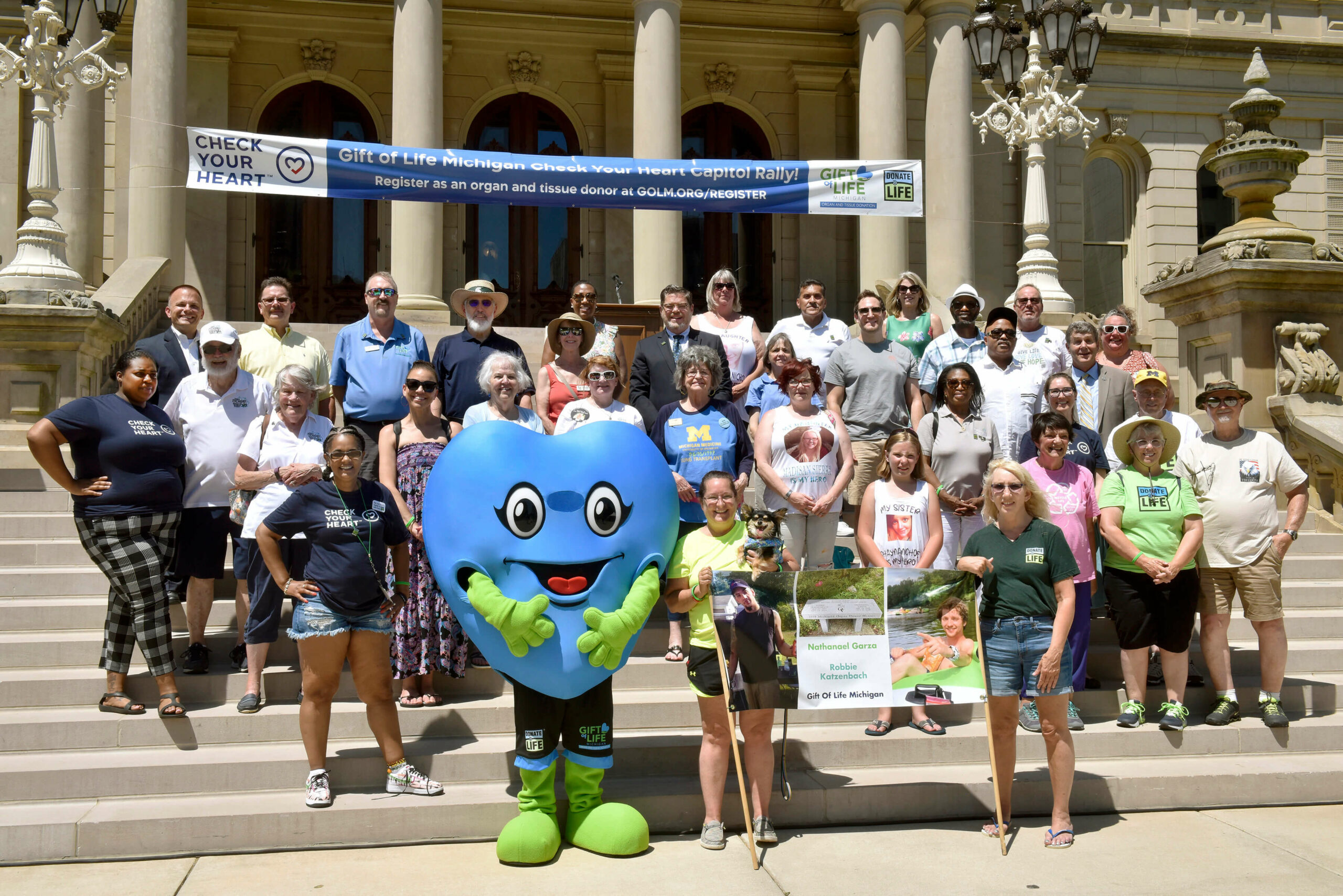 A group of organ and tissue donation supporters gathered on the Capitol steps with Hartley (blue heart mascot) under a Check Your Heart banner