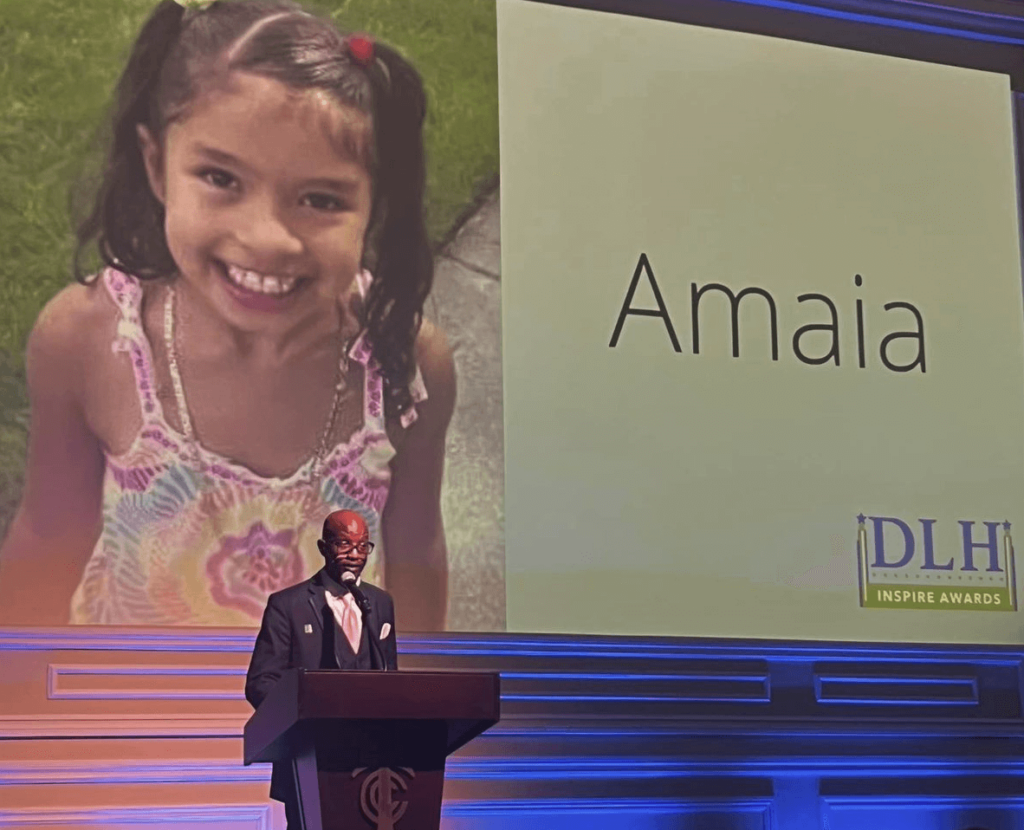 Man at podium in front of extra large screen showing photo of a little girl and her name, Amaia