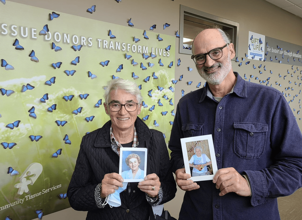 Man and woman standing in front of butterfly mural holding up photos of an elderly woman
