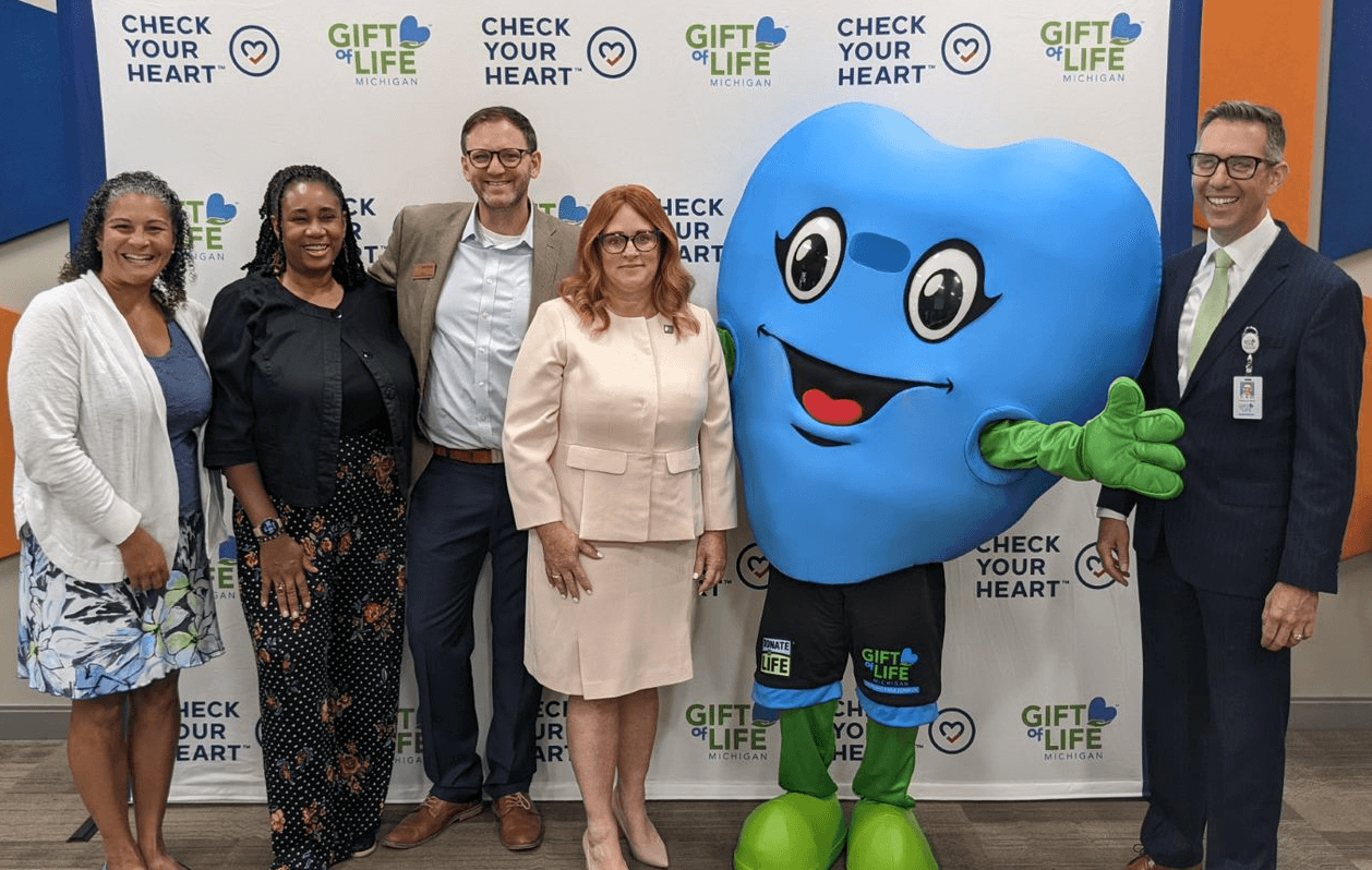 A group of legislators and executives gather in front of a logo "step and repeat" with Hartley, a large blue heart-shaped mascot