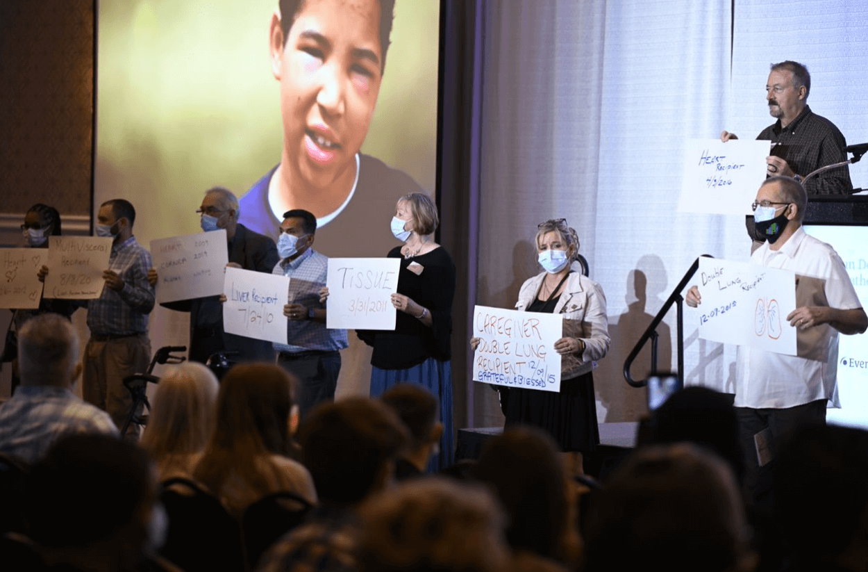 Group of transplant recipients holding 'Thank You' signs toward the donor family audience, in front of a stage and a video playing on a large screen