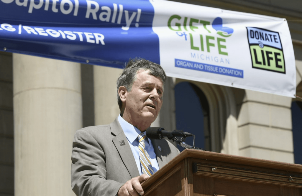 Senator Kevin Daly stands at a podium in front of a Check Your Heart banner on the Michigan Capitol steps