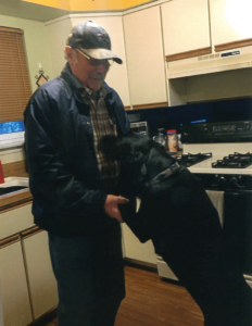 Black lab on its hind legs, with front legs being held by an older man in his kitchen