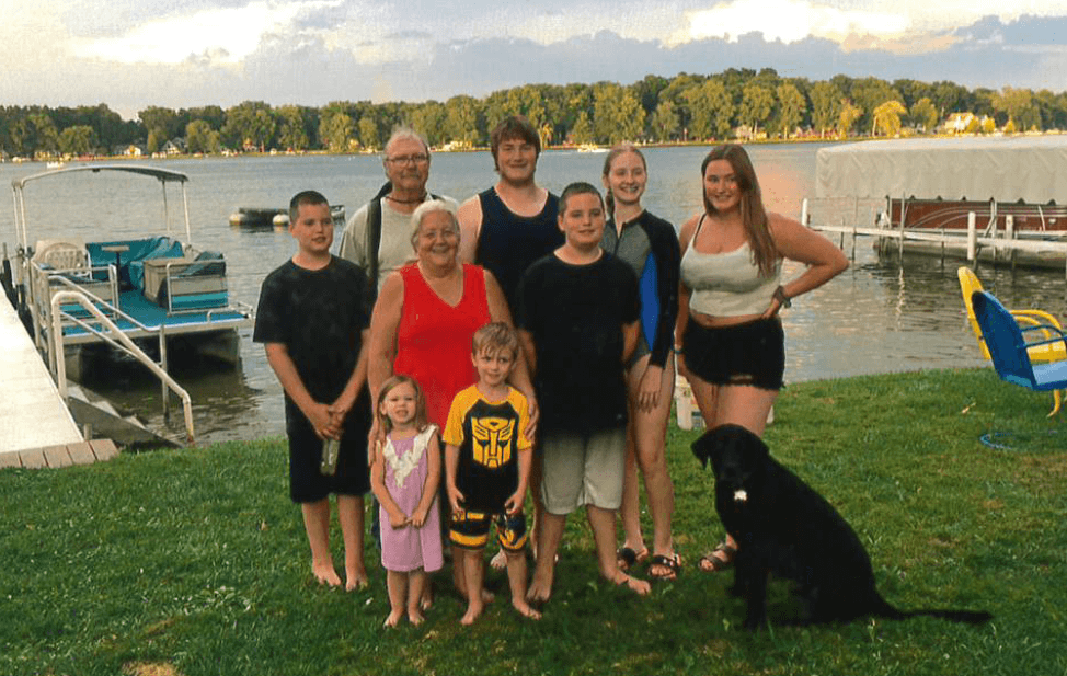 multi-generational family photo on the grass in front of a lake with a boat at the dock behind them