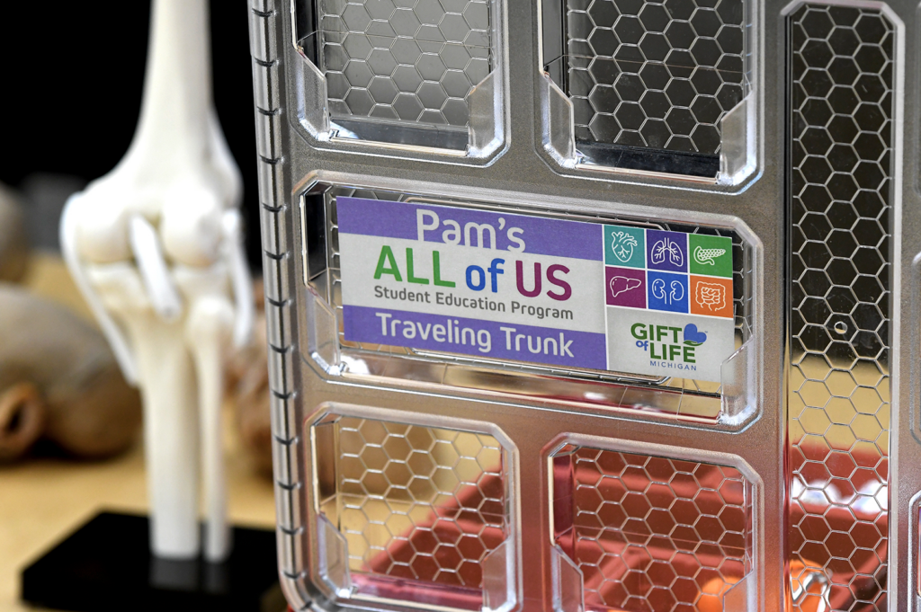 Red plastic case with plastic bone sample, with a cover that reads "Pam's All of Us Student Education Traveling Trunk" with the Gift of Life Michigan logo