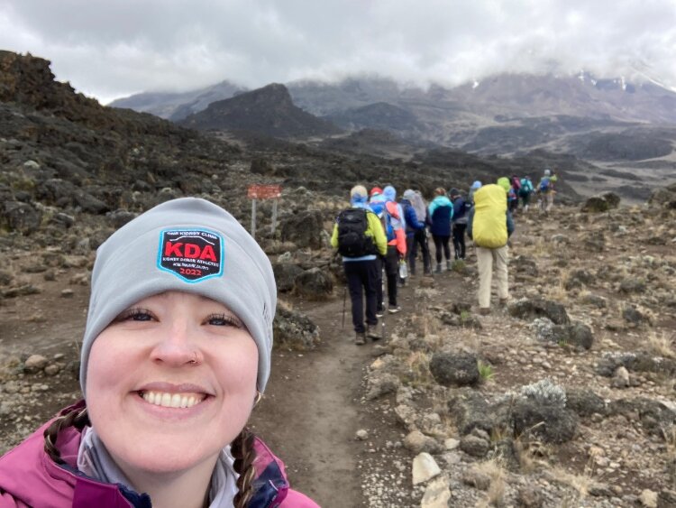 Emily Polet-Monterosso smiling for a selfie-style photo while hiking toward a snow-capped mountain with other hikers.