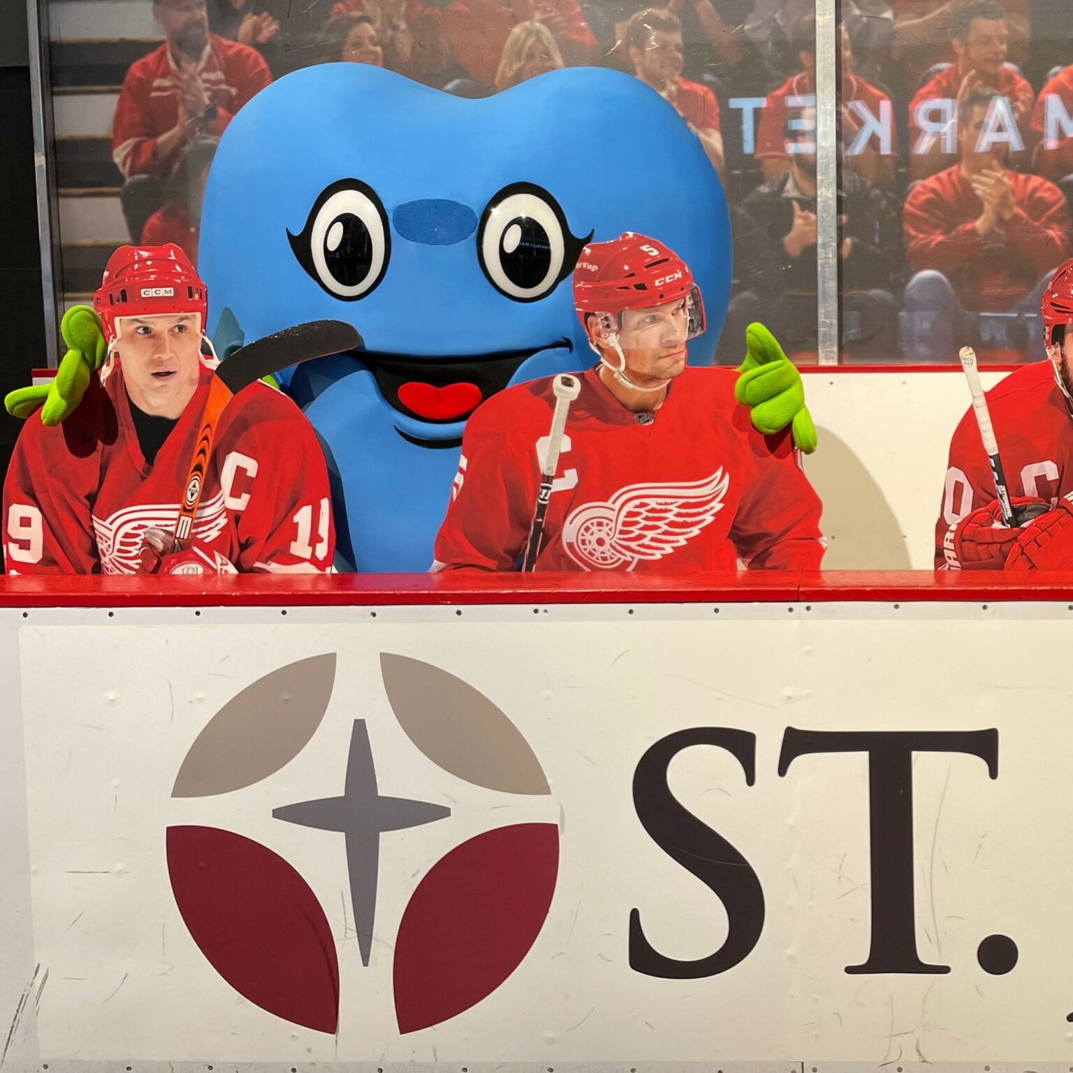 Hartley, a large blue heart-shaped mascot, in the box with a couple of Red Wings in uniform.