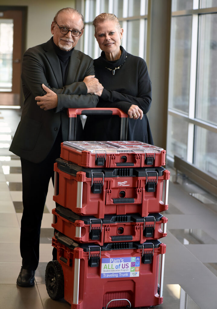 Pam and Krishna Sawhney standing with the red "Pam's All of Us Traveling Trunks"