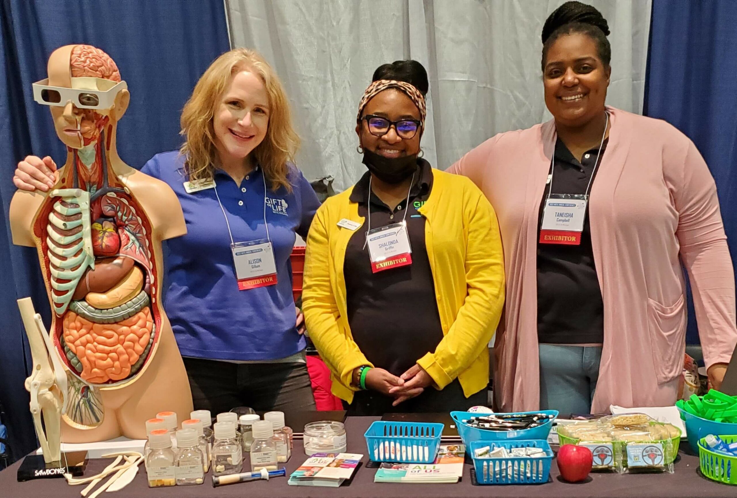 Gift of Life community relations coordinators standing behind a table with their All of Us high school outreach supplies