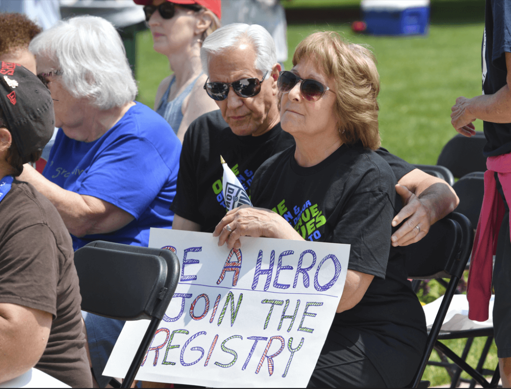 A woman seated in front of the Michigan Capitol steps with a sign reading "Be a Hero, Join the Registry."