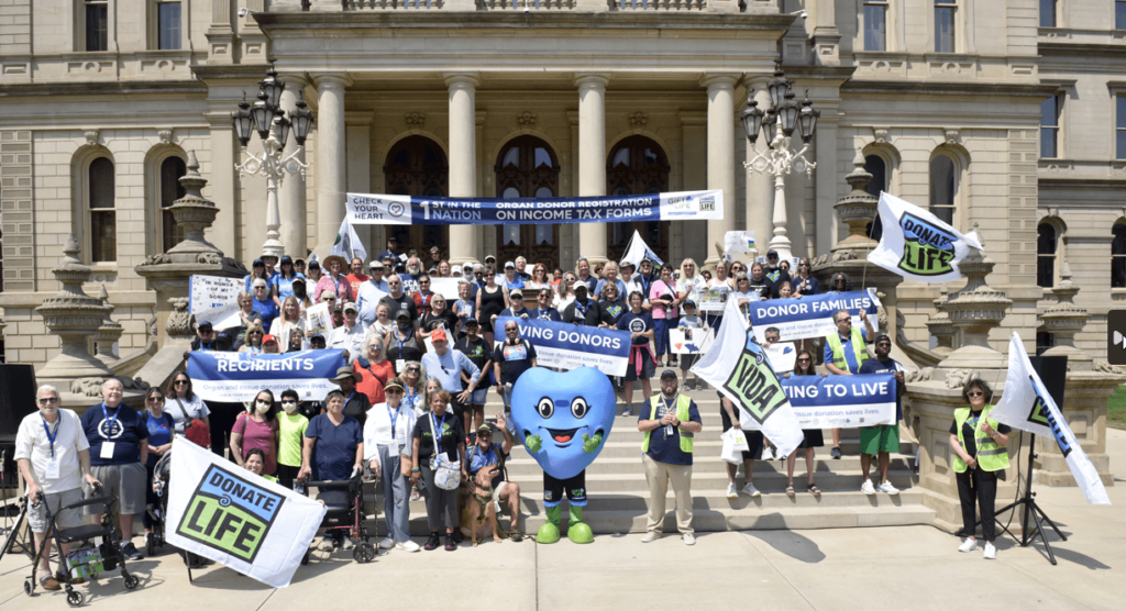 A large crowd gathered on the Michigan Capitol steps (including mascot Hartley T. Heart) holding Donate Life flags and banners for donor families, living donors, transplant recipients and those waiting to live.