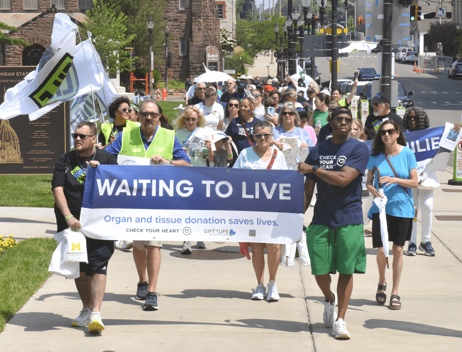 A group walks down the sidewalk in front of the Michigan Capitol holding a banner reading "Waiting to Live."