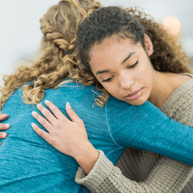 Two women hugging, the one facing the camera has a somber look on her face.