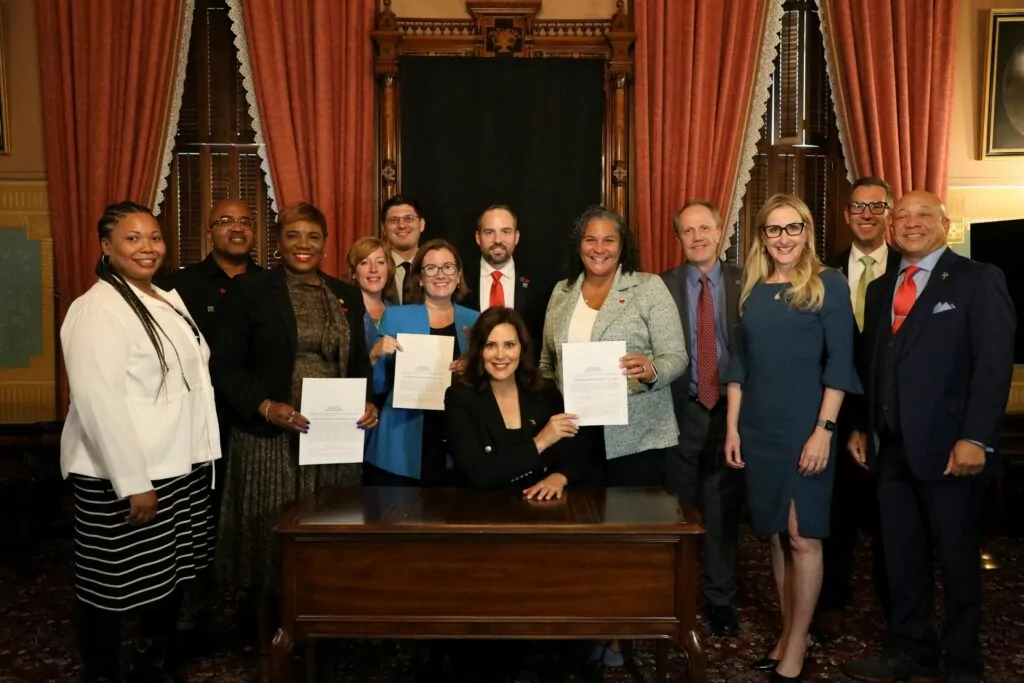 A group of Gift of Life staff, transplant recipients and legislators gathers around Governor Whitmer, who is seated at her desk and holding up the signed Check Your Heart Act