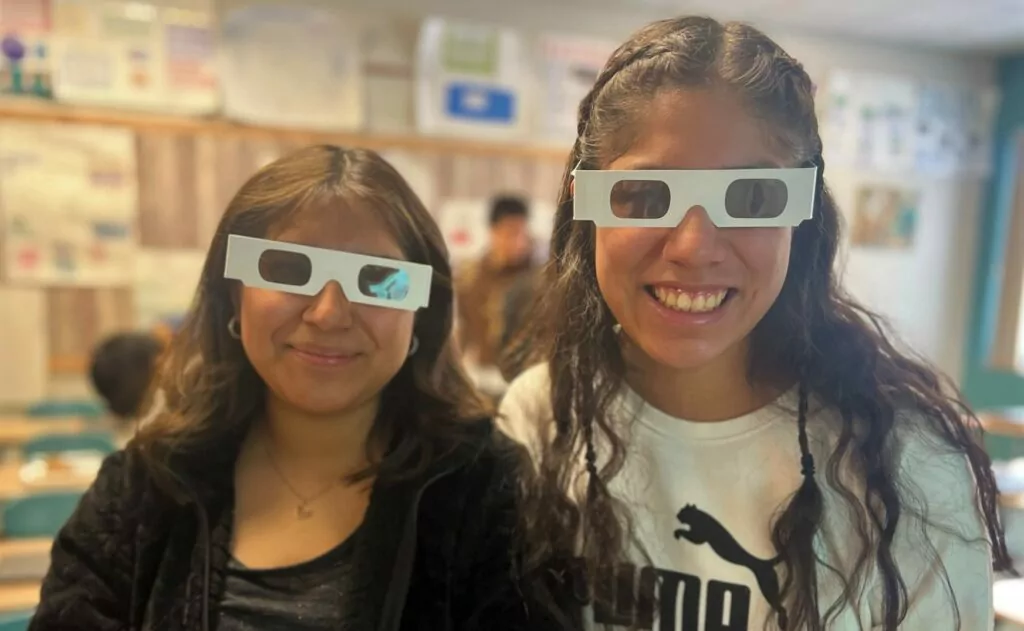 Two teenage girls wearing disposable glasses that mimic the need for a cornea transplant.