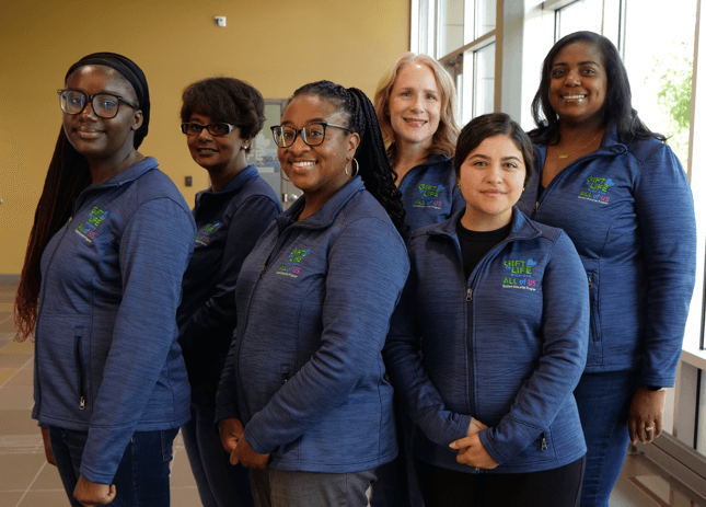 Six women wearing "All of Us" branded jackets