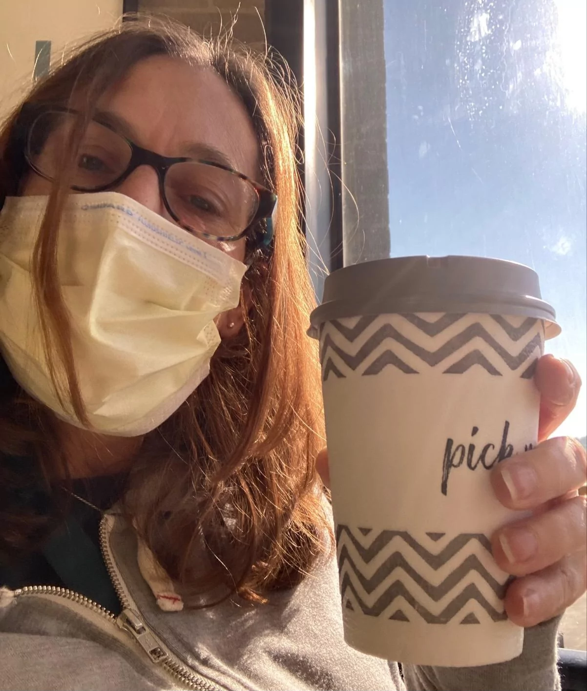 Sherry holding a cup of coffee, while wearing a mask in front of the window of her hospital room
