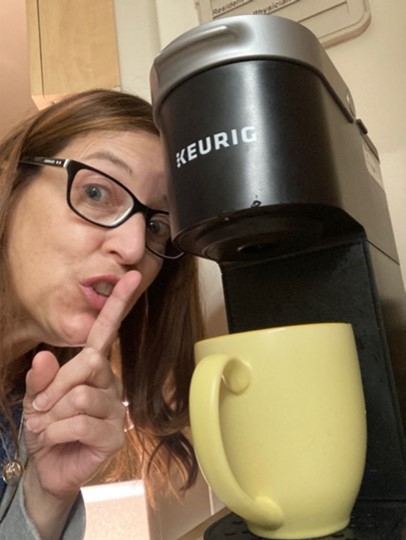 Sherry with her finger over her lips in a "shush" sign, next to a Keurig coffee maker and mug.
