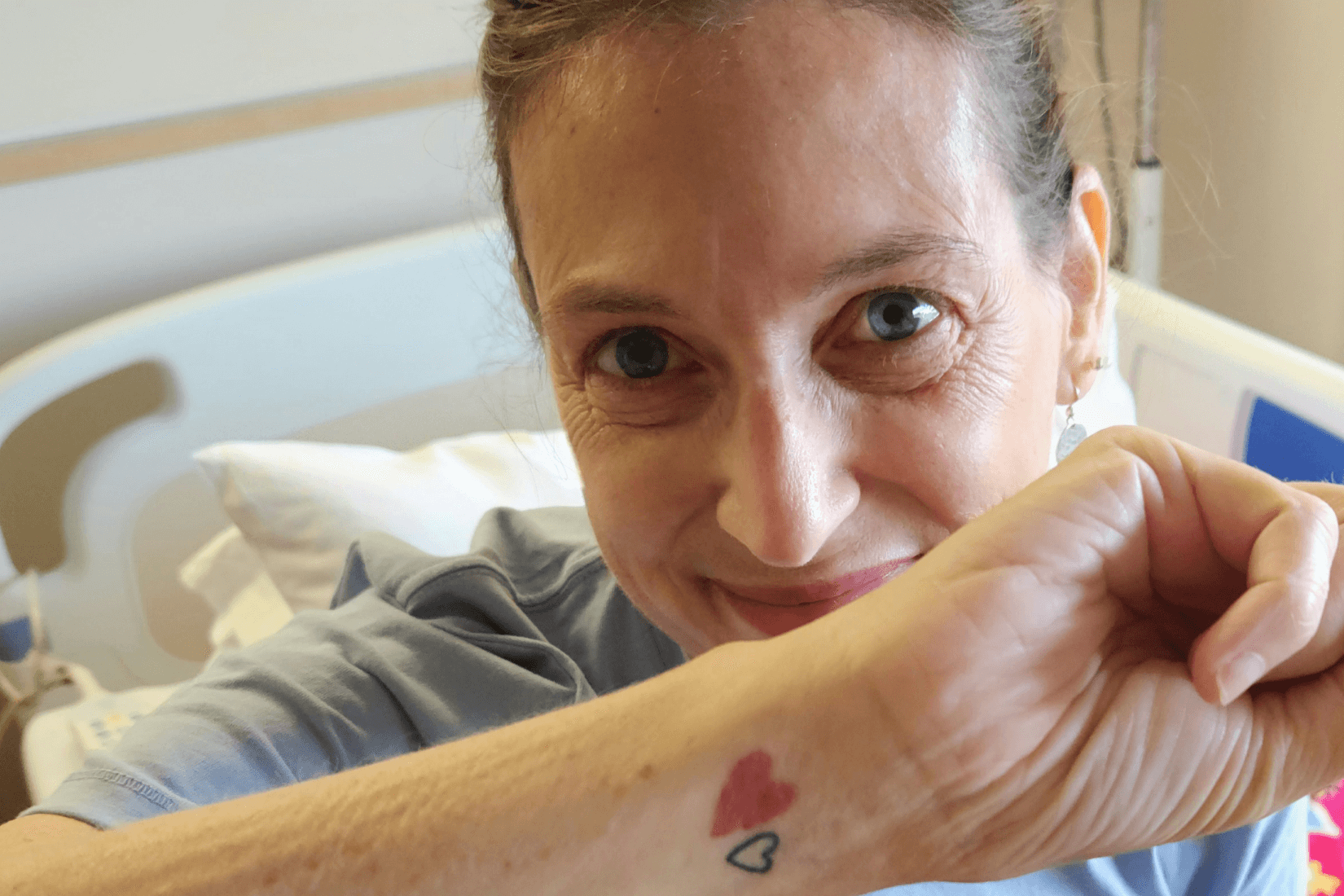 Sherry Johnson, with hair pulled back right, sits on her bed in her hospital room. Her forearm is facing the camera so we can see her red heart tattoo.