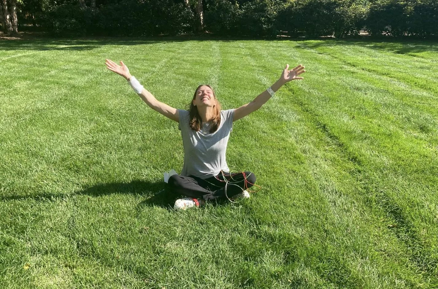 Sherry Johnson, outside on a large, green lawn with trees behind her, arms outstretched and face up taking in the sun