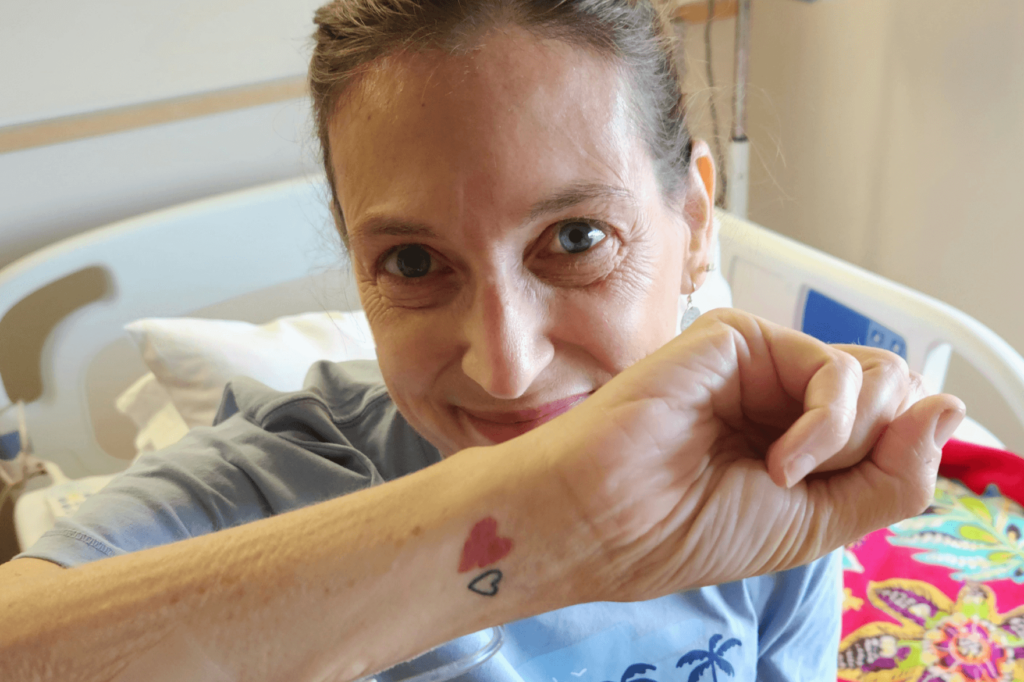 Sherry Johnson, with hair pulled back right, sits on her bed in her hospital room while she waits for a second heart and kidney transplant. Her forearm is facing the camera so we can see her tattoo, representing her original heart and her first transplanted heart.