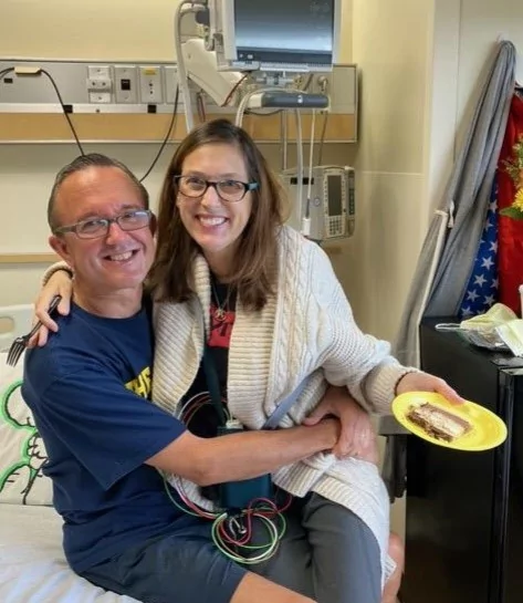 Sherry sitting on Tim's lap on her hospital bed, holding a piece of cake on a paper plate.