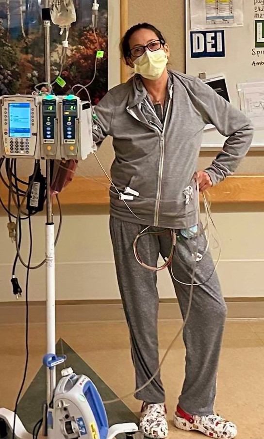 Sherry Johnson in her street clothes and crocs, one hand on her hip the other holding her IV pole as she walks around the unit post-heart/kidney transplant