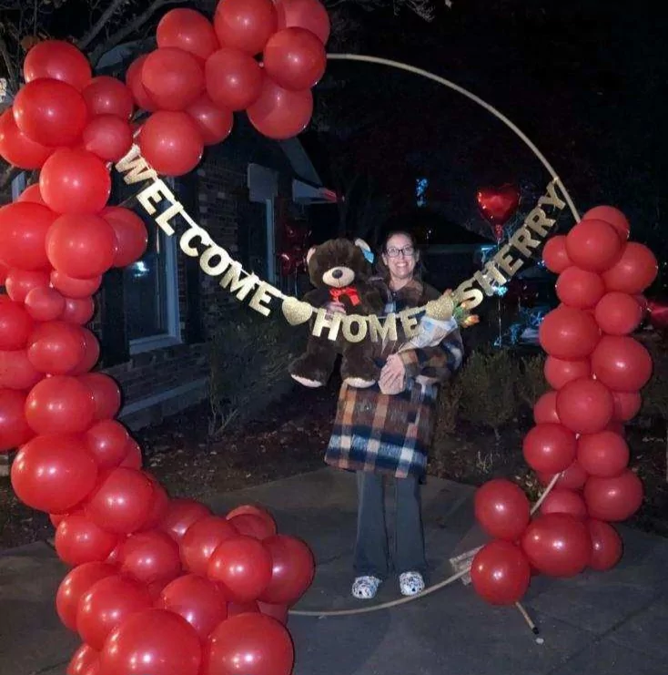 Sherry Johnson stands behind a large red balloon display with the words "Welcome Home Sherry" hanging, holding a giant teddy bear, after returning home from her heart and kidney transplant.