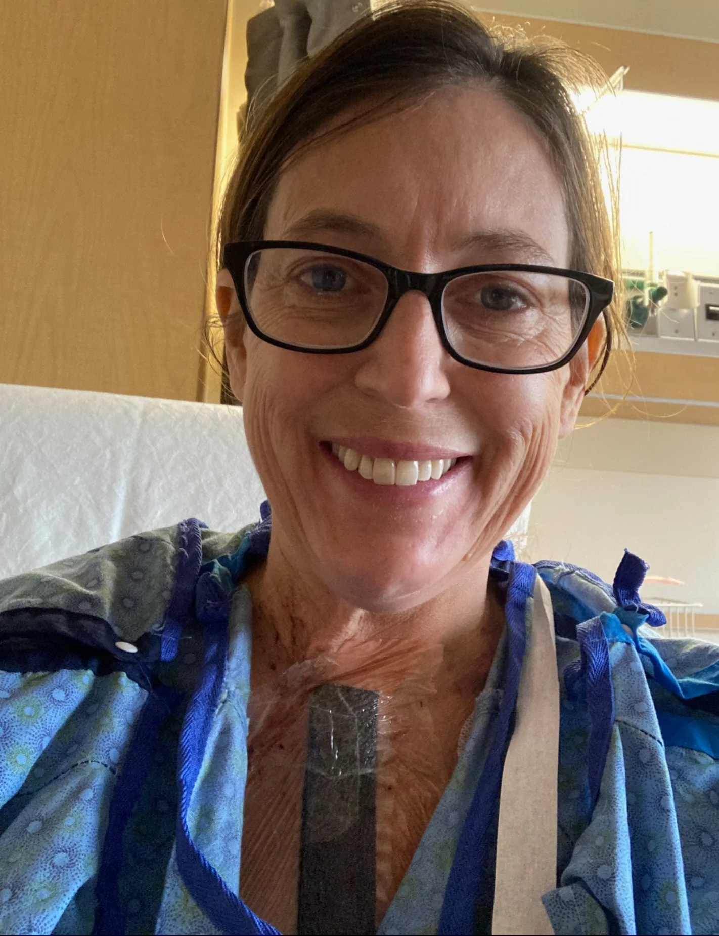 Sherry Johnson sitting up on her hospital bed, in a gown with surgical tape down her sternum, and smiling.